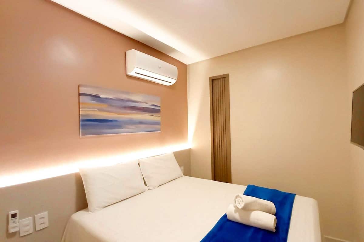A hotel room with a bed, an air conditioner, a painting, a television, and a door.