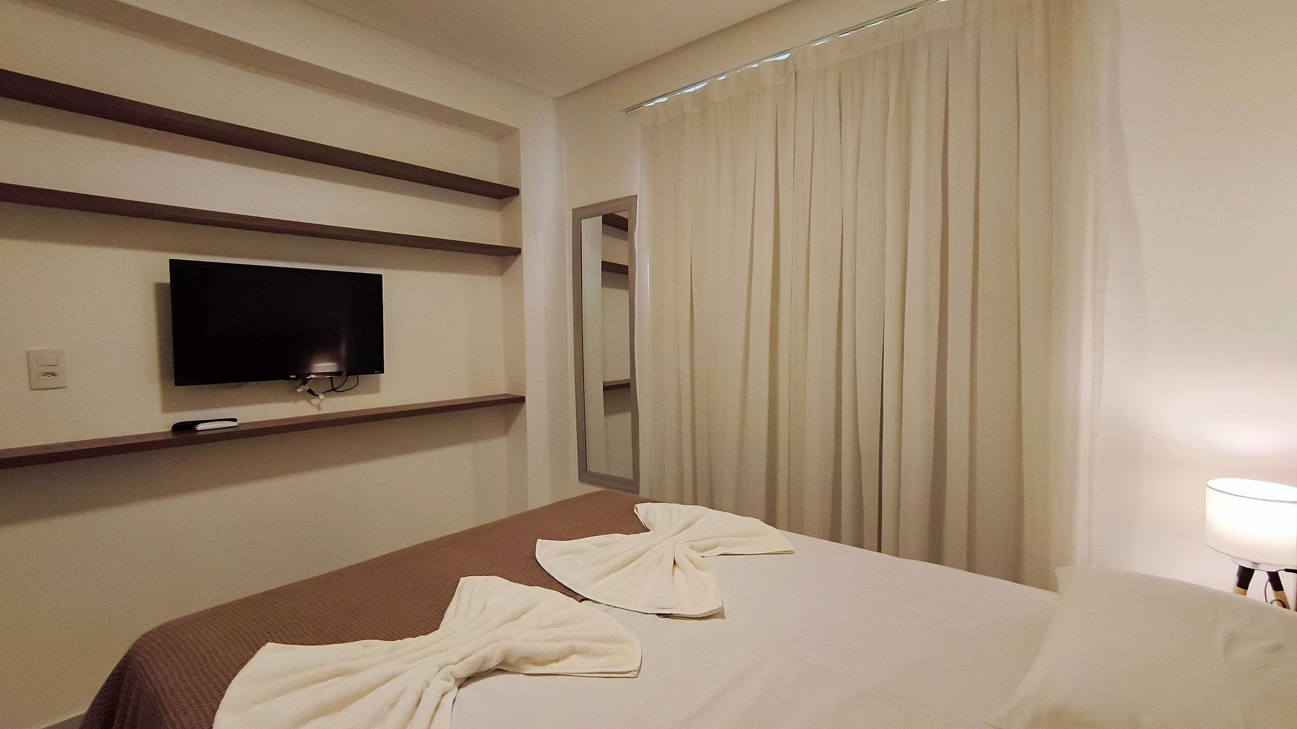 A hotel room with a bed, a TV mounted on the wall, a mirror, and a window with curtains.