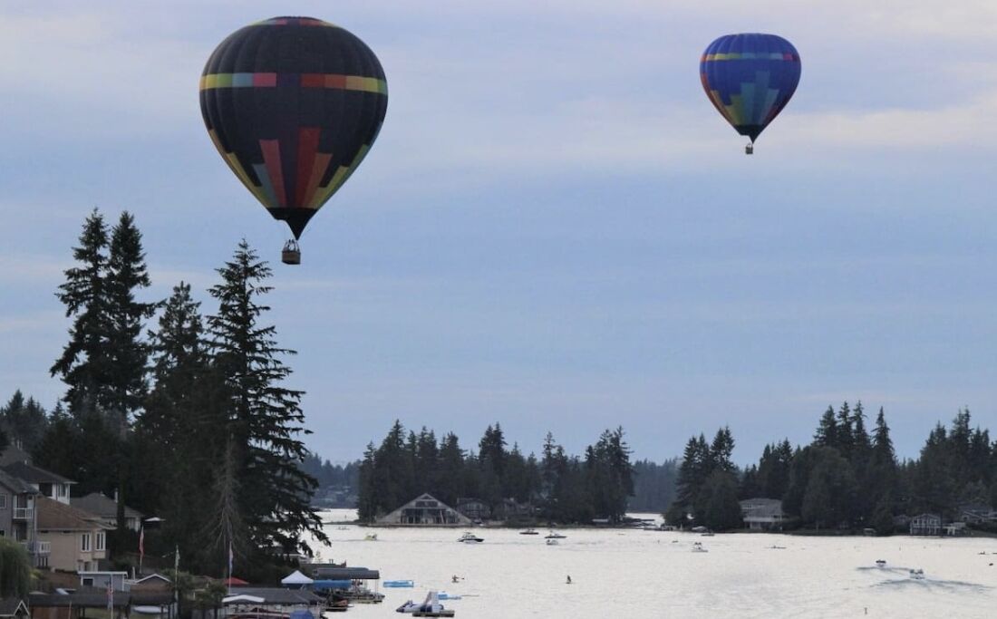 Two hot air balloons flying over a waterfront area with houses and boats.
