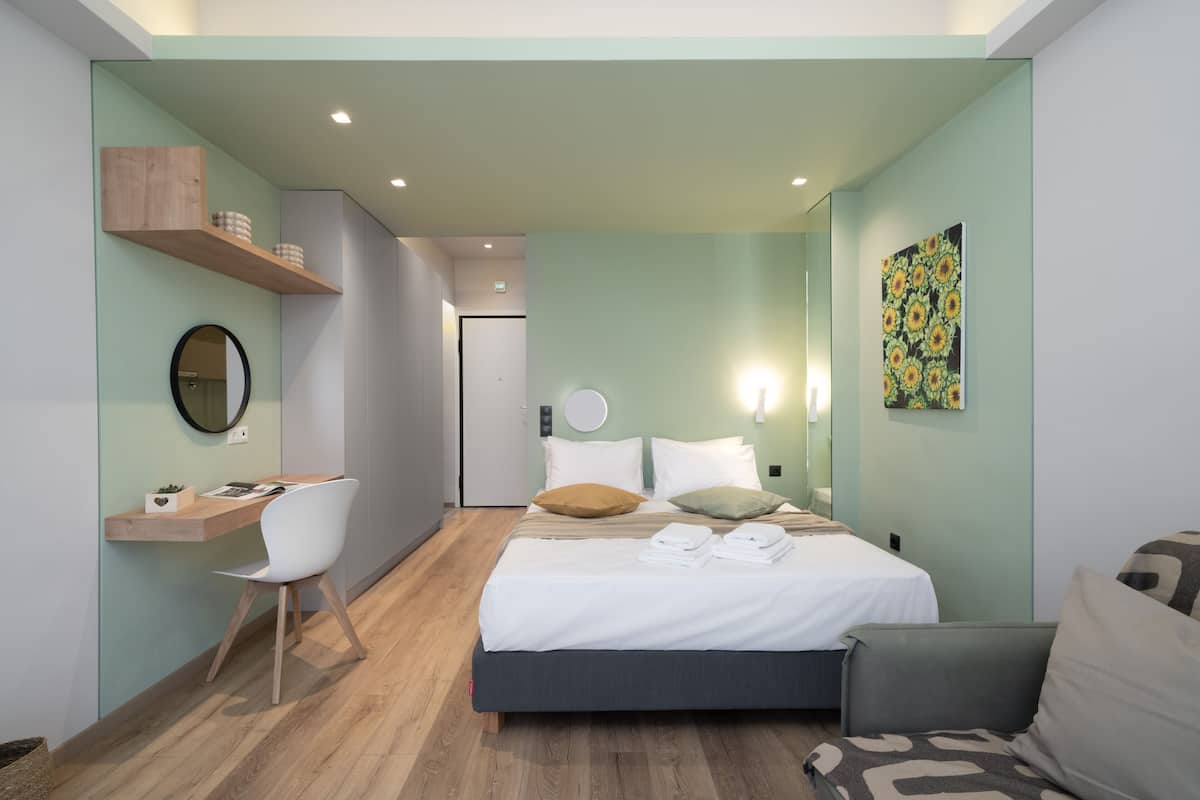 A modern hotel room with a bed, a desk, and a chair. There is a wall-mounted shelf with decorative items.