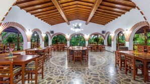 A spacious dining area with wooden tables and chairs, surrounded by arched windows with greenery visible outside.