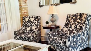 A living room with two patterned armchairs, a glass coffee table, a side table with a lamp, and a mirror on the wall. - Azalea Suite at The Mockingbird Inn (Ocean Springs)