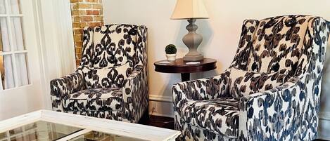 A living room with two patterned armchairs, a glass coffee table, a side table with a lamp, and a mirror on the wall.