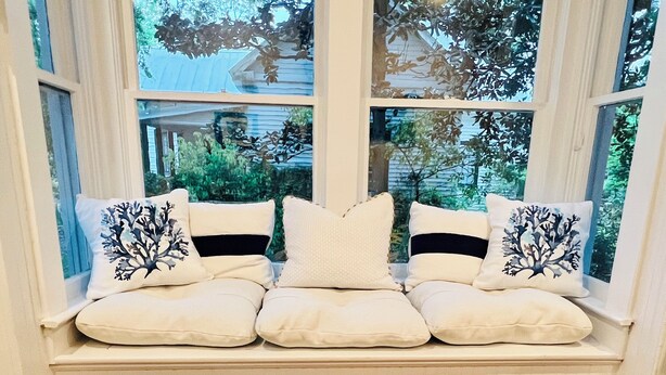 A white bench with cushions and decorative pillows against a window with a view of greenery and a house. - Azalea Suite at The Mockingbird Inn (Ocean Springs)