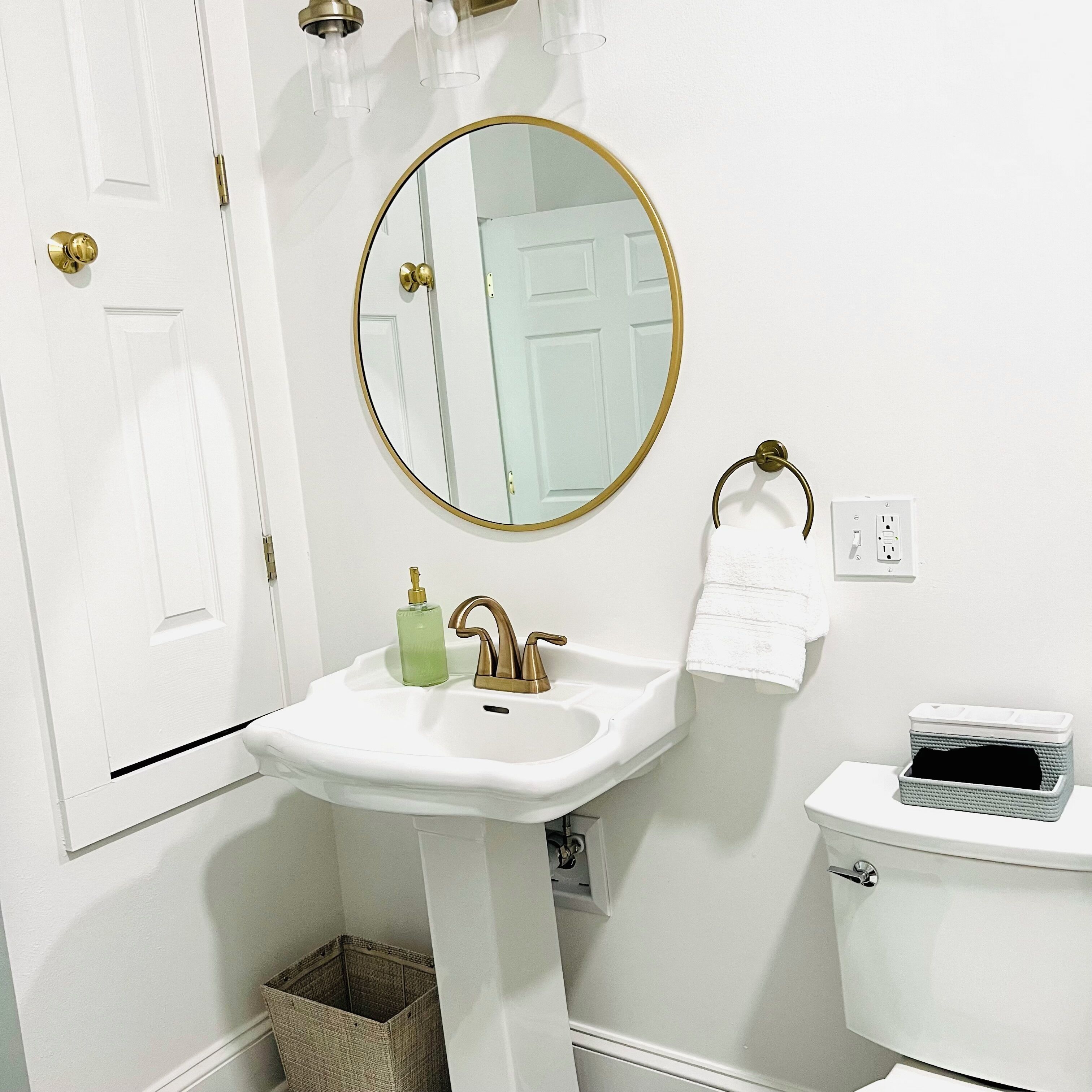 Un baño con un lavabo blanco, un espejo con marco dorado, un inodoro blanco y una cesta debajo del lavabo.