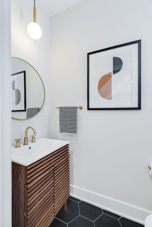 A modern bathroom with a white sink, gold faucets, a round mirror, and a framed abstract artwork on the wall.