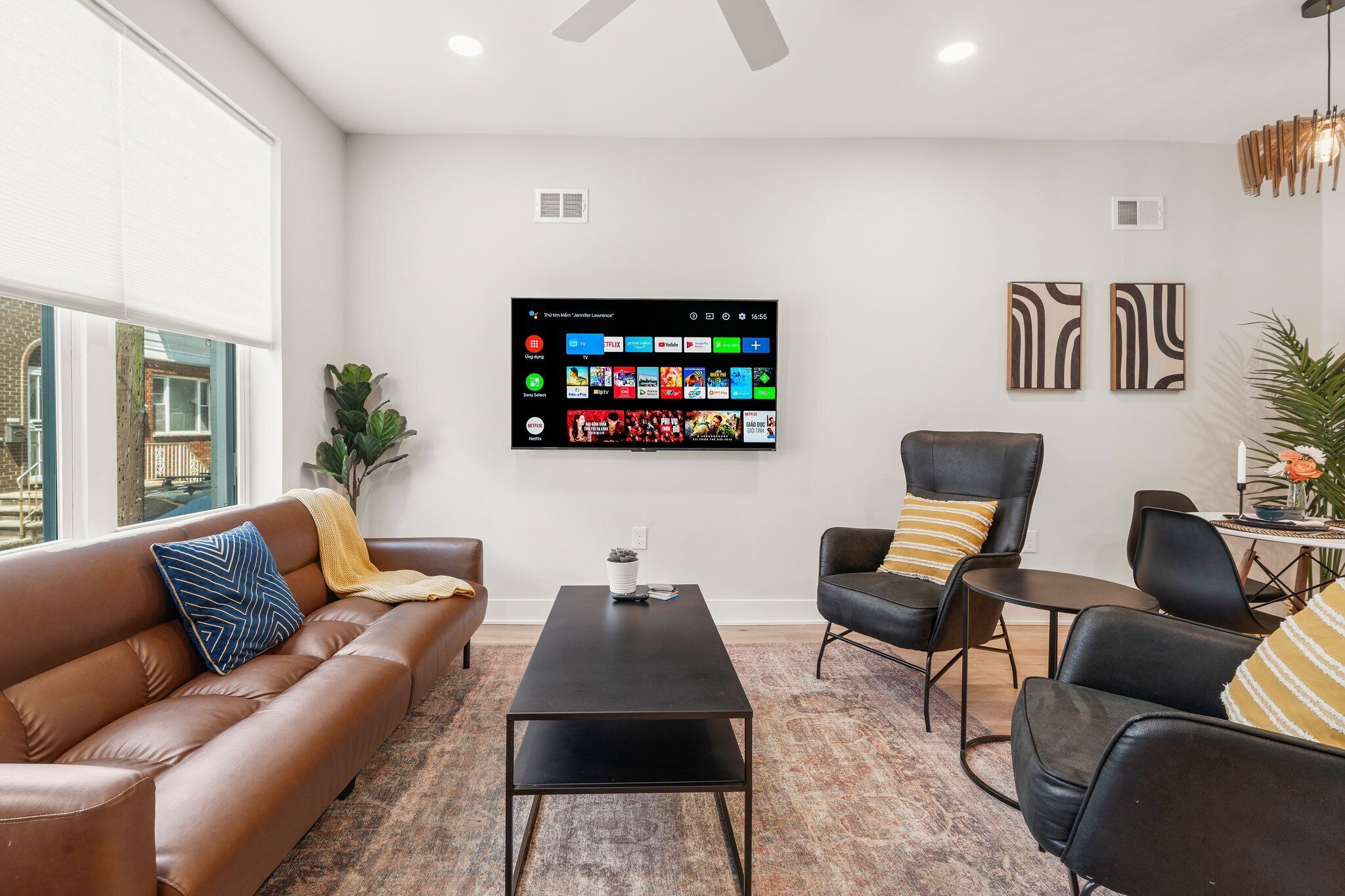 A modern living room with a leather sofa, a coffee table, and two armchairs. A flat-screen TV mounted on the wall displays various streaming services.