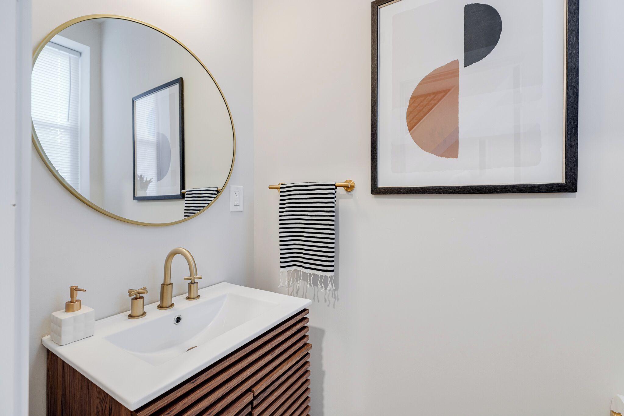 Een badkamer met een witte wastafel, gouden kraanwerk, een ronde spiegel en een gekaderd abstract schilderij aan de muur.