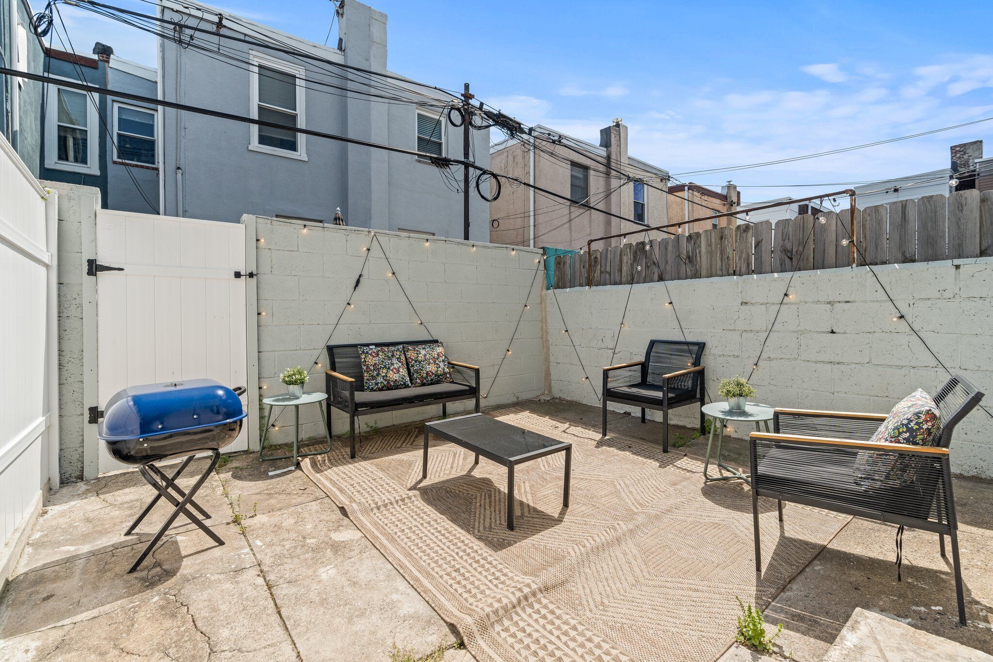 A backyard with a barbecue grill, two black metal chairs, a small round table, and a sofa with colorful cushions.