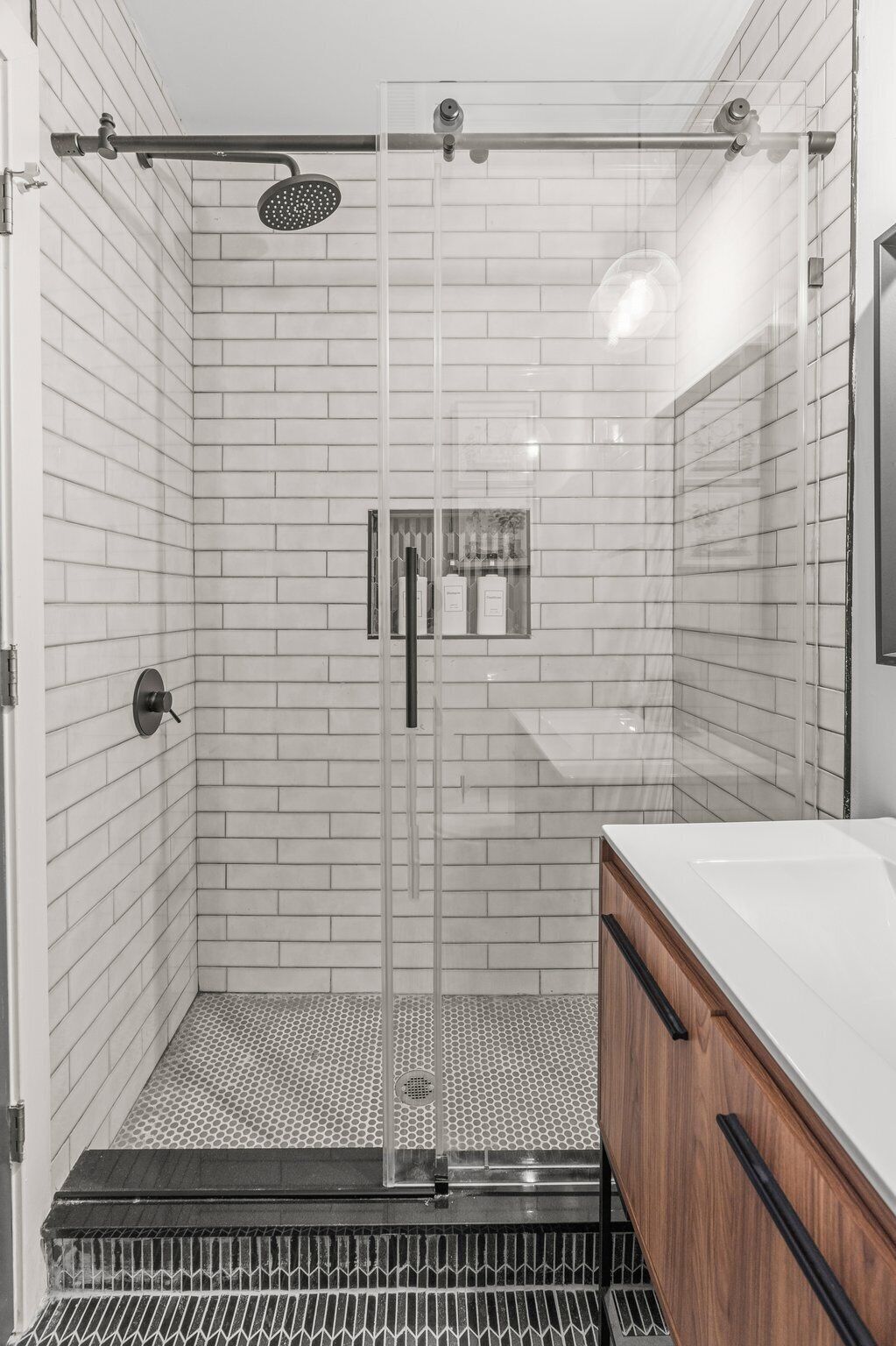 Une salle de bain moderne avec une douche à parois de verre, des murs recouverts de carreaux blancs, un meuble-lavabo en bois et une grille en métal noir en bas.