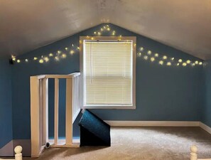 A room with a sloped ceiling, a window with blinds, a wooden crib, and string lights.
