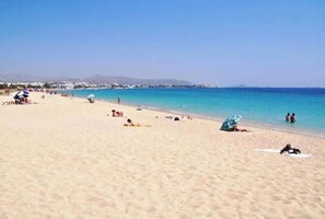 A beach with people sunbathing and swimming, clear blue water, and a distant mountain range.