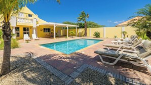 Un espace piscine avec des chaises longues, un coin salon abrité et un bâtiment jaune doté d’un balcon.