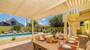 Un espace extérieur abrité, aménagé pour les repas, avec une piscine, des murs jaunes et un jardin tropical.