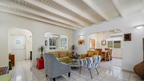Un vaste espace de vie comprenant un canapé, des fauteuils et une table basse. On y trouve également un coin repas avec une table et des chaises. La pièce est dotée d’un sol carrelé et de murs blancs.