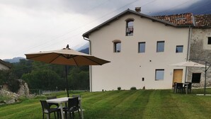 Une cour arrière verdoyante avec une table et des chaises sous un parasol, un bâtiment doté de nombreuses fenêtres et un mur en pierre.