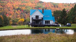 A house with a blue roof and white walls, situated next to a pond with a forested hillside in the background.
