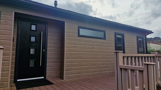 Una terraza de madera con una puerta y ventanas negras, un pequeño porche y un cielo nublado.