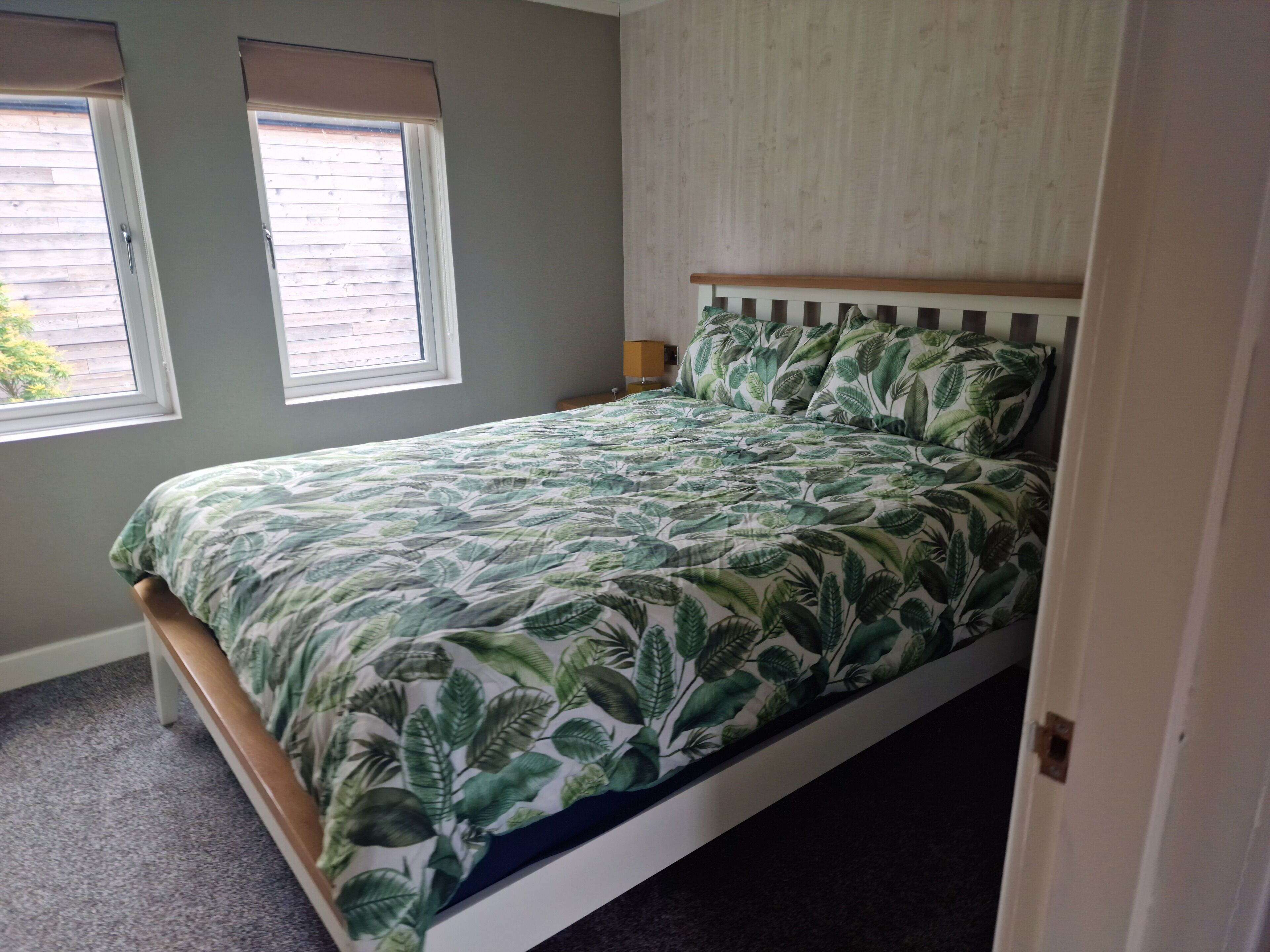 A single-bed room with a wooden headboard, a green leaf-patterned bedspread, two windows with blinds, and a light-colored wall.
