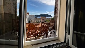 A balcony with wooden chairs and a table, overlooking a coastal area with buildings and a hill.