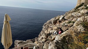 A rocky cliff with a stone building, a chair, and a table with a beige umbrella.