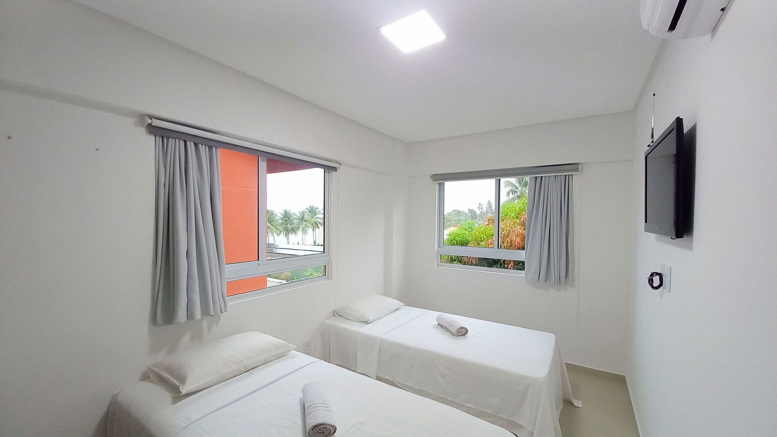 A hotel room with two single beds, a TV mounted on the wall, and windows with curtains.