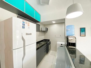 A compact kitchen with a refrigerator, microwave, stove, and water dispenser. - Lindos apartamentos no Porto Cayman (Tamandaré)