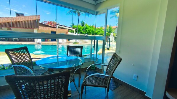 A balcony with a glass enclosure overlooking a swimming pool, wicker chairs, and a glass table.