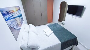 A hotel room with a bed, pillows, a blanket, a TV, and a painting of a sunset over the ocean. - Lindos apartamentos no Porto Cayman (Tamandaré)