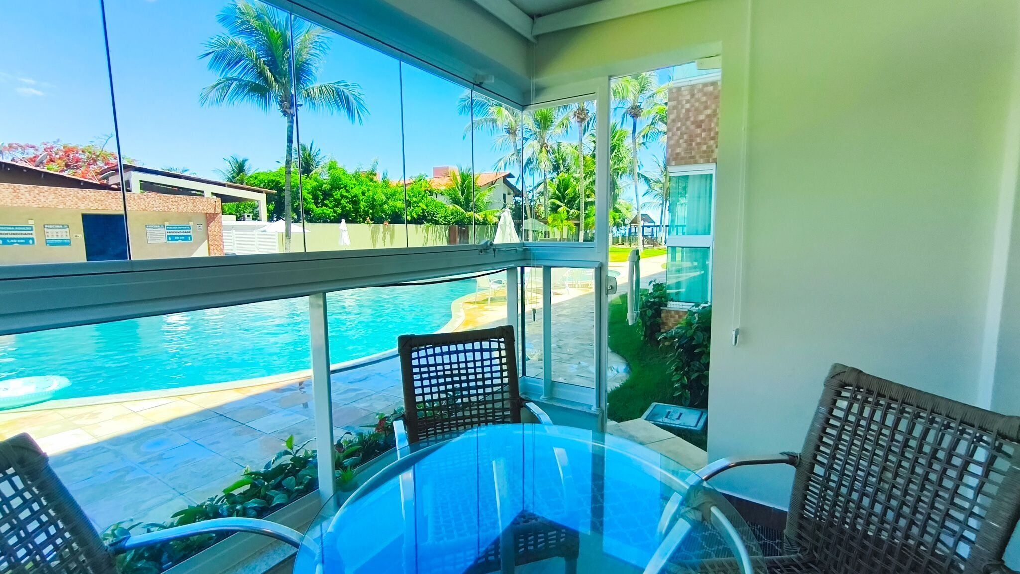 A glass-enclosed balcony with a pool view, wicker chairs, and a glass table.