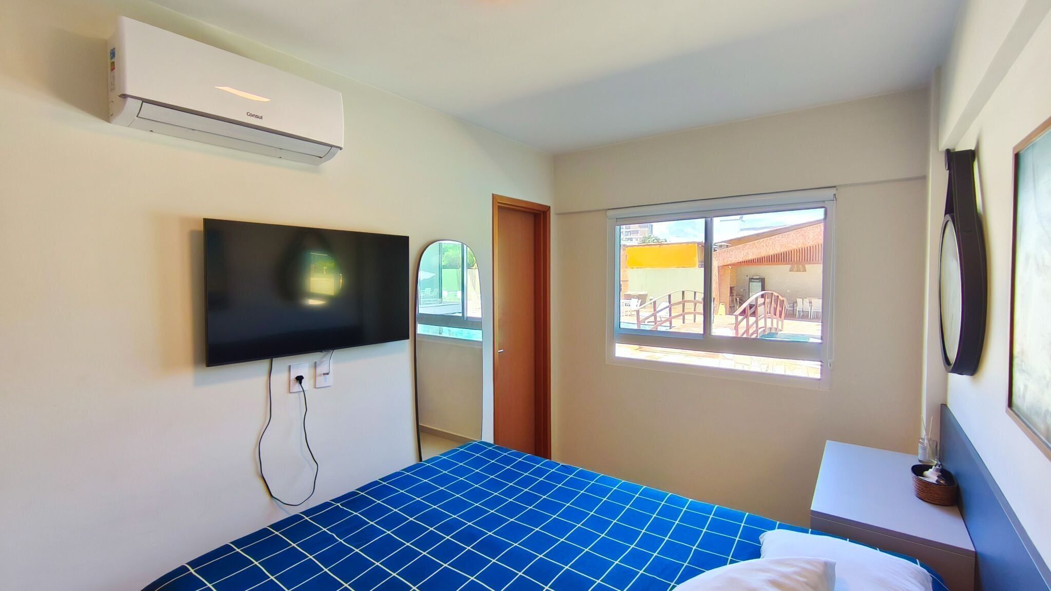 A hotel room with a bed, a mounted TV, an air conditioner, a window with a view of the outside, and a small table with a lamp.