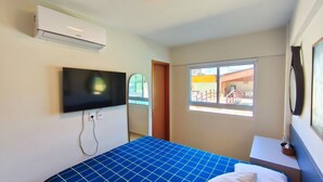 A hotel room with a bed, a mounted TV, an air conditioner, a window with a view of the outside, and a small table with a lamp. - Lindos apartamentos no Porto Cayman (Tamandaré)