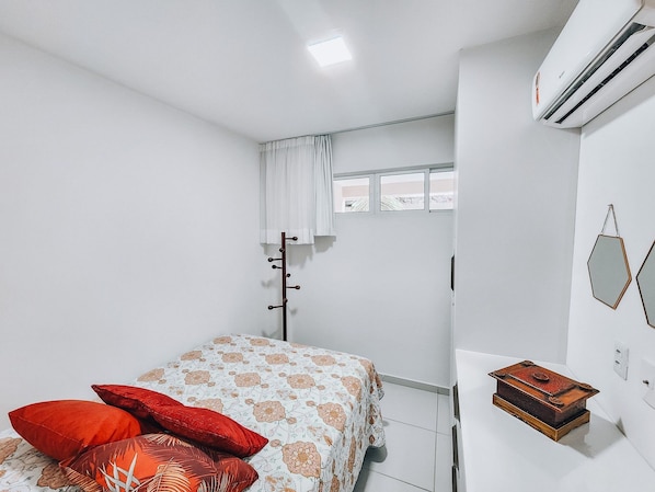 Un dormitorio con una cama, una ventana con cortinas, un aire acondicionado y un pequeño cofre de madera.