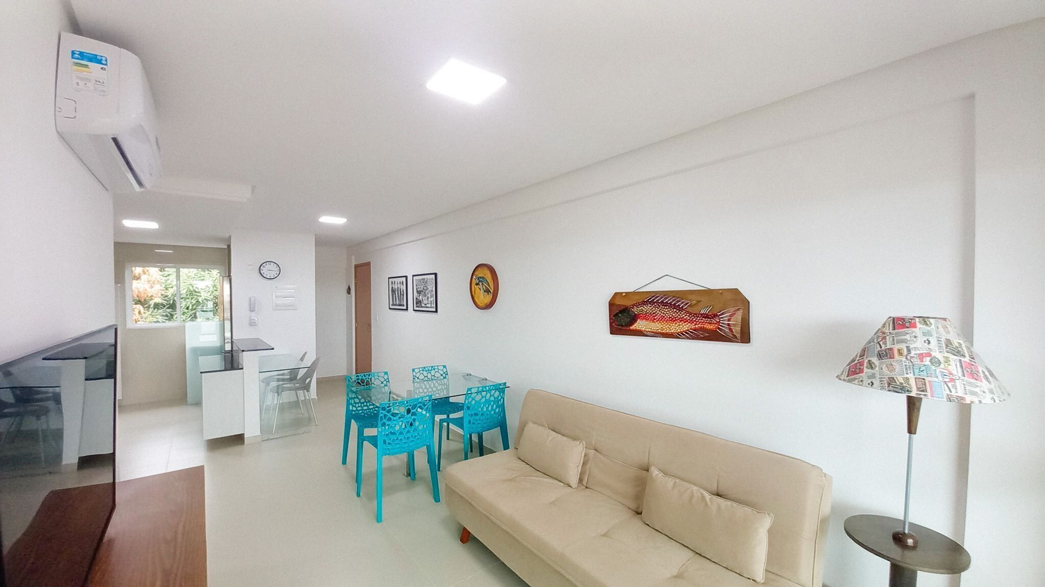 A modern living room with a beige sofa, a glass dining table with blue chairs, a wall-mounted fish decoration, and a lamp with a patterned lampshade.
