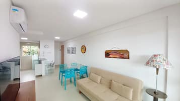 Uma sala de estar moderna com um sofá bege, uma mesa de jantar de vidro com cadeiras azuis, um enfeite de peixe na parede e um abajur com cúpula estampada.
