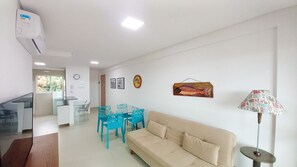 A modern living room with a beige sofa, a glass dining table with blue chairs, a wall-mounted fish decoration, and a lamp with a patterned lampshade. - Lindos apartamentos no Porto Cayman (Tamandaré)