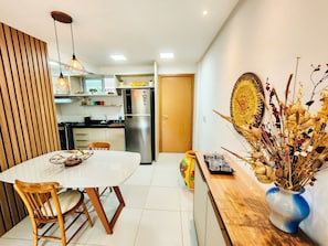 A modern kitchen with a refrigerator, stove, and dining area with a table and chairs. - Lindos apartamentos no Porto Cayman (Tamandaré)