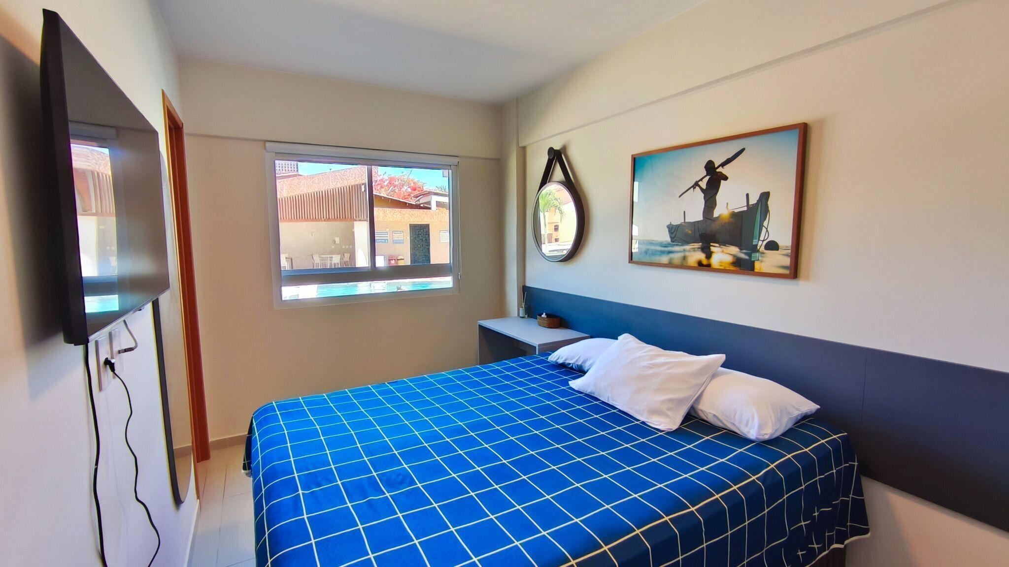 A hotel room with a bed, a TV, a window, and a painting on the wall.