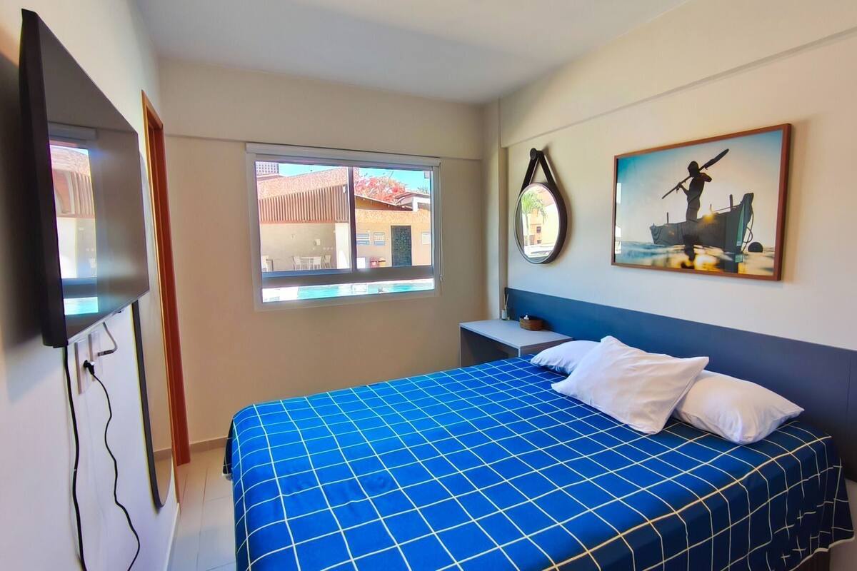 A hotel room with a bed, a TV, a window, and a painting on the wall.
