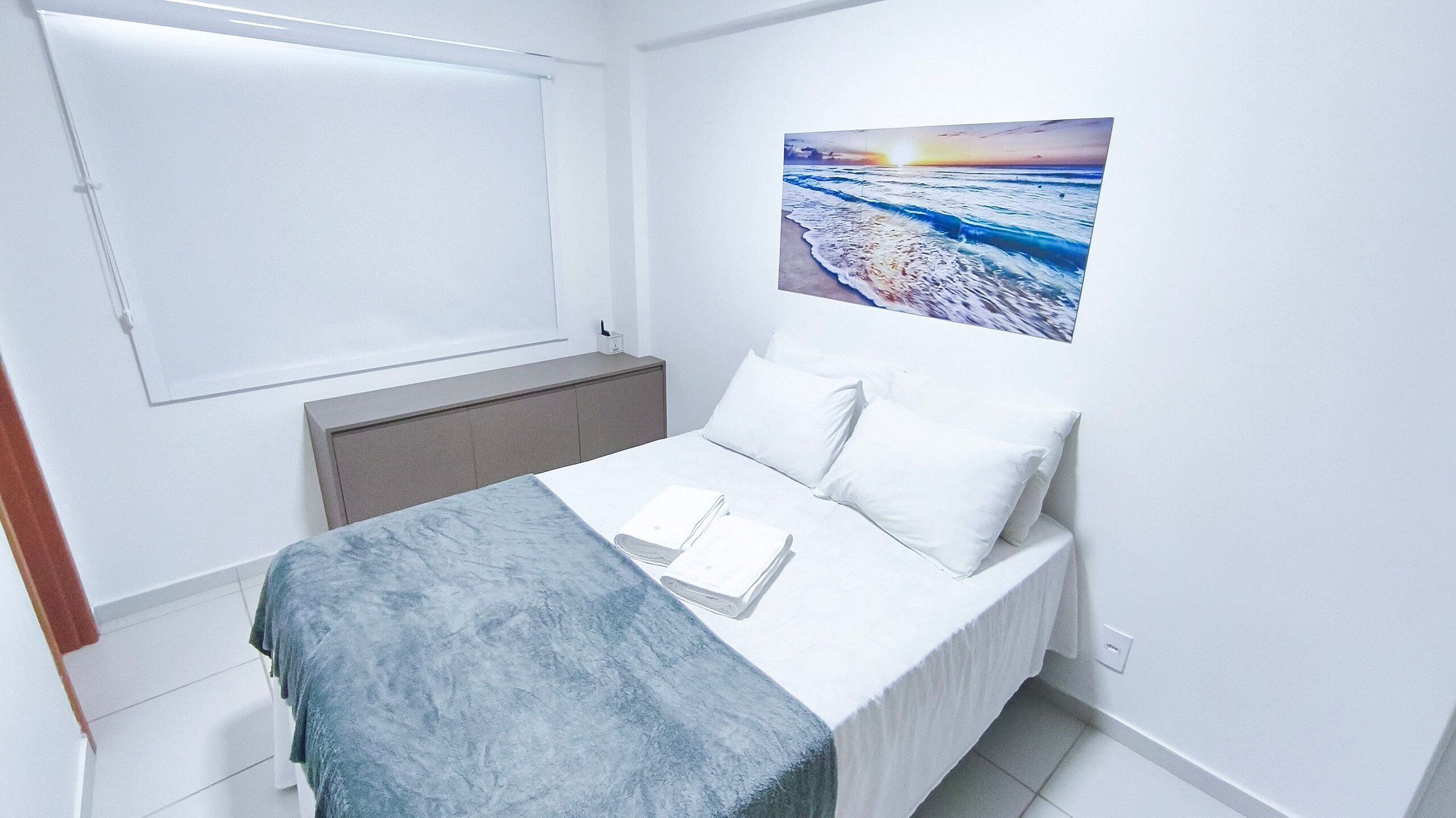 A bedroom with a bed, a window, a painting of a beach scene, and a bedside table with a book and a lamp.