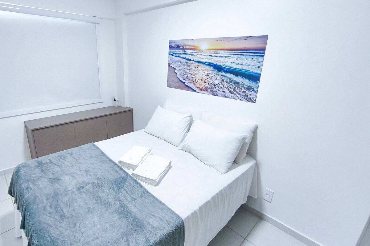 A bedroom with a bed, a window, a painting of a beach scene, and a bedside table with a book and a lamp.