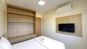 A hotel room with a bed, built-in wooden shelving, a mounted TV, and an air conditioning unit.