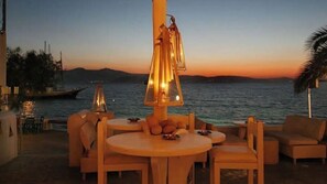 Un espacio al aire libre con mesas y sillas de madera redondas, un accesorio de iluminación decorativo, y vistas al mar y montañas distantes al atardecer.