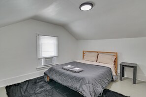 A bedroom with a sloped ceiling, a bed with grey bedding, a window with blinds, a bedside table, and a rug.