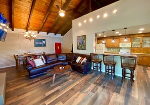 A spacious living room with a large L-shaped sectional sofa, a wooden coffee table, and a dining area with a wooden table and chairs. The kitchen features wooden cabinets and a bar area with stools. - Lakefront, 50 Guests, Paradise Estates, Reunions   - , Weddings, Work Retreats (Lakehead)