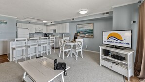 A modern living room with a dining area, kitchenette, and a television displaying 'OC Beachfront Rentals'.