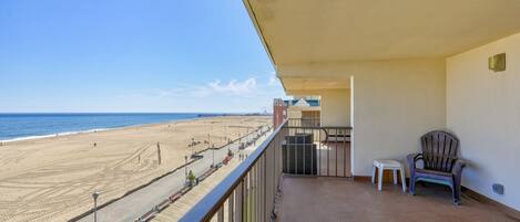 A balcony with a view of the beach, a chair, and a small table.