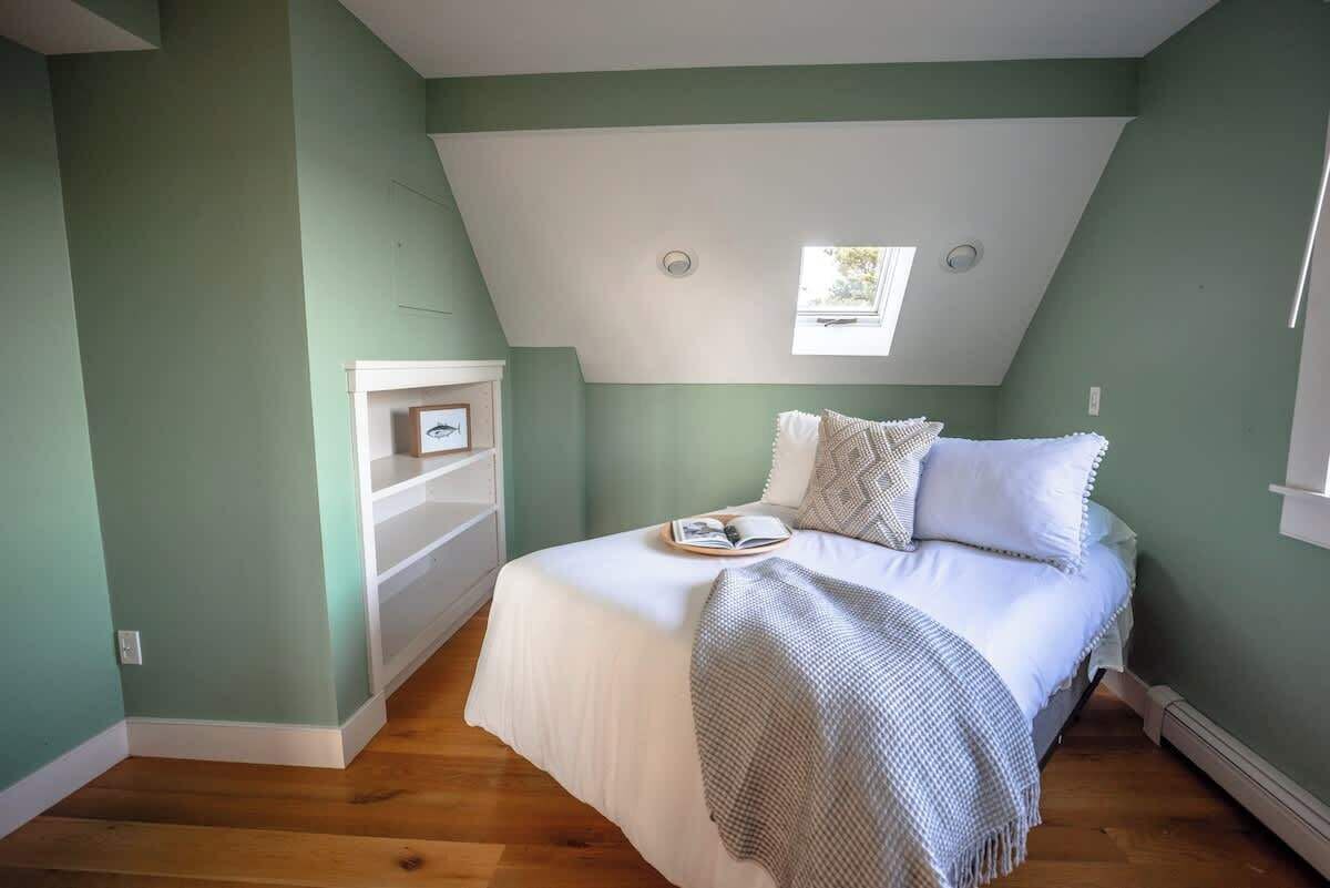 Un dormitorio con techo inclinado, una cama con ropa de cama blanca, un ventanal pequeño y un estante blanco.
