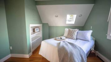 Un dormitorio con techo inclinado, una cama con ropa de cama blanca, un ventanal pequeño y un estante blanco.