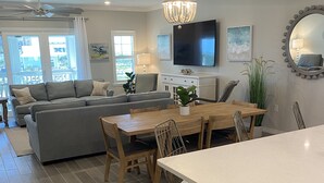 Un salon moderne avec un canapé gris, une table à manger en bois et des chaises. On y trouve un téléviseur fixé au mur, un lustre et un grand miroir.